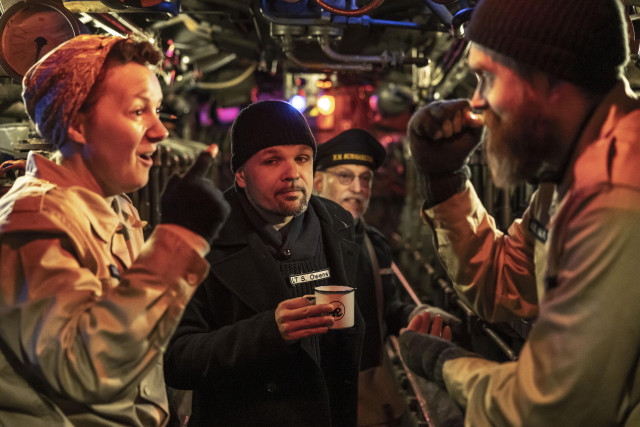 Vier Personen in engem Raum, der Kulisse eines U-Boots. Rote Beleuchtung. Vorne scheinen ein bärtiger Mann und eine Frau mit Kopftuch heftig zu diskutieren. Dahinter schauen zwei Männer zu. Einer hält eine weiße Emaille-Tasse. Alle vier tragen Seefahrer-Uniformen.