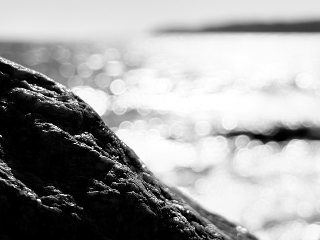 Black and white photo of a black rock by the sea, with the water and a distant island out of focus.