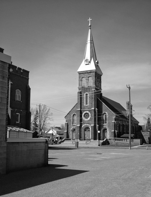 A black and white image of a church with a tall shiny metal covered steeple. A large cross is seen at the very top of the steeple and large stairs lead up to the bulding. Another building can be partially seen on the left edge of the photo.