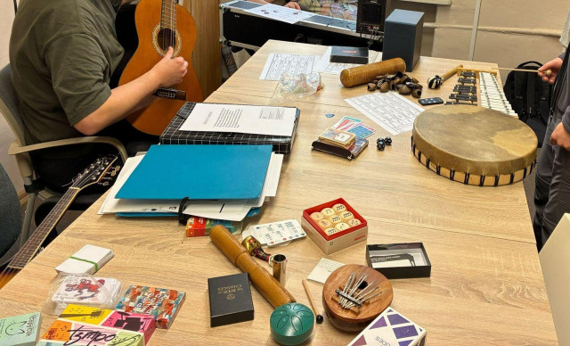 A large table with small instruments and gaming equipment. Around the table three people with a guitar, modular synth, and percussion.