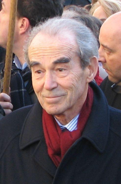 Image de Robert Badinter. Il porte un manteau noir, une chemise bleue claire et une écharpe rouge. Il est au milieu d’une foule. La photo semble avoir été prise à l’extérieur pendant la journée.