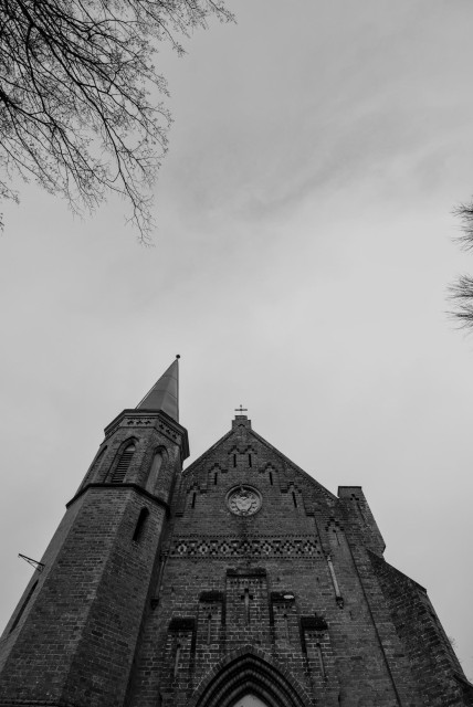 Kirchturm mit spitzem Dach und Uhr an der Fassade, von unten fotografiert, mit kahlen Baumzweigen am Bildrand