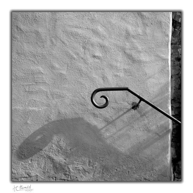 A square black-and-white photograph with a white border, showing a white, roughly plastered house wall. A curved metal banister rises into the frame from the right. Halfway up, it bends horizontally and ends in a small spiral. On the far right, a small section of a rough stone wall can be seen, and the banister casts a shadow downwards to the left. With a little imagination, this shadow looks like the head of the Loch Ness monster.