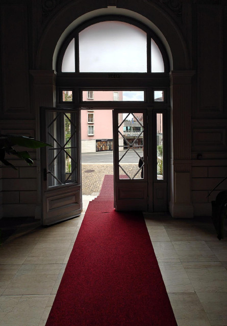 A photo looking out through an arched entryway with glass doors. A borrow red or burgundy carpet extends through the open doorway to the paved street outside. 