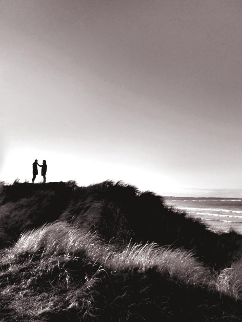 On a sun-drenched, high dune on Strandhill beach in Sligo, Ireland, two people stand in the distance as dark silhouettes, facing each other. The person on the right is touching the person opposite with one hand, at face or shoulder height. 
On the horizon to the right, the ocean can be seen breaking against the shore beneath a vast, cloudless sky. 

Auf einer sonnenbeschienenen hohen Düne am Strand von Strandhill, Sligo, Irland stehen in der Distanz zwei Menschen als dunkle Silhouetten, einander zugewandt. Die Person rechts berührt mit einer Hand die gegenüberstehende Person in Höhe des Gesichts oder der Schulter. 
Am Horizont ist rechts der heranbrandende Ozean unter einem weiten, wolkenllosen Himmel zu sehen.