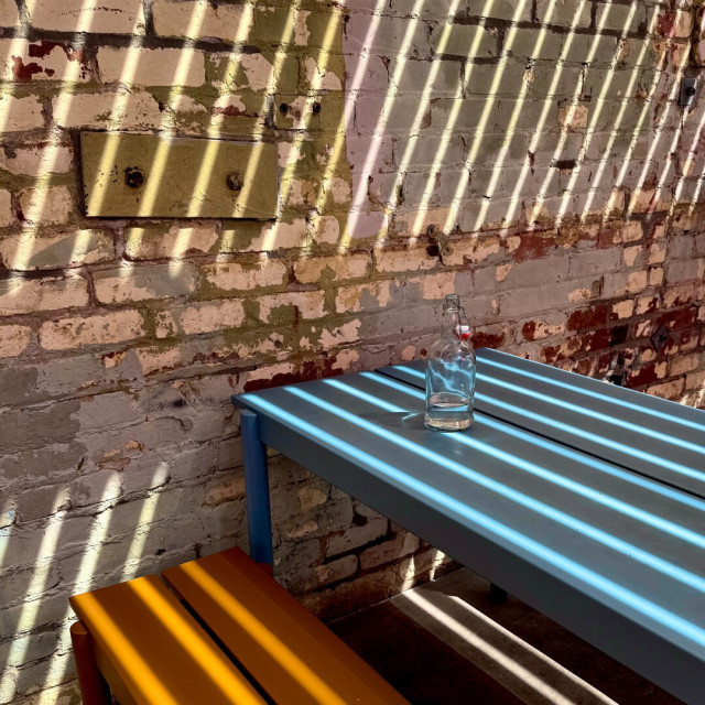 Blue metal picnic table with an orange bench against a brick wall with a lot of paint wear. A slatted awning off-camera is producing angled shadows below.