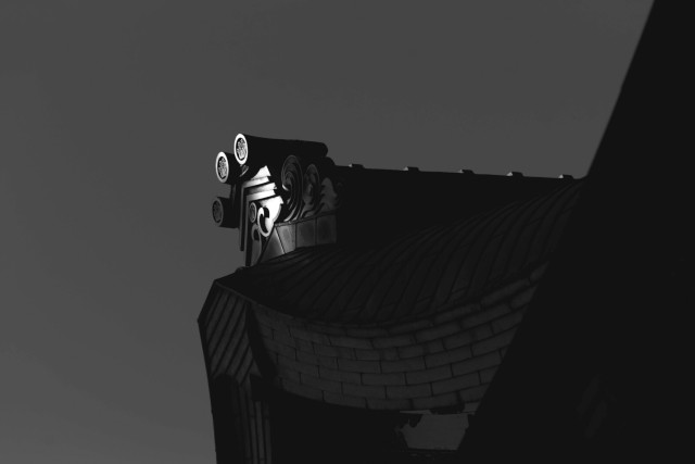 Black and white photo of a tile, a stylised house shape topped with three scrolls, that catches the sun while the rest of the flared roof on which it sits remains in shadow.  I think this is an 'onigawara' on the Chokushi-Mon in Kew Gardens.