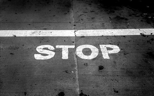 A worn “STOP” marking painted on wet pavement, scattered with leaves and small debris, split by a faint seam down the center.