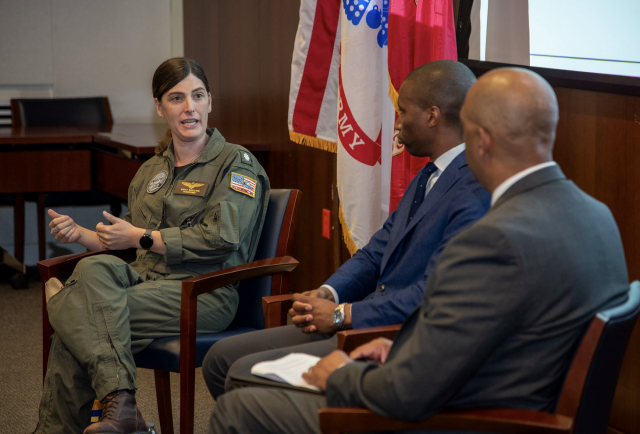 Emily Schilling in her flight suit, talks to two men in non-flight suits. She is speaking and they are listening.