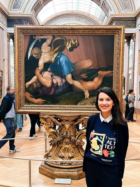 A vertical photo showing yours truly (a Caucasian woman with long brown hair) standing in front of the painting "The Battle of David and Goliath" in the Grande Gallerie of the Louvre Museum. I am wearing a navy cardigan, navy wide leg pants and the Mastodon t-shirt "I write alt text" 