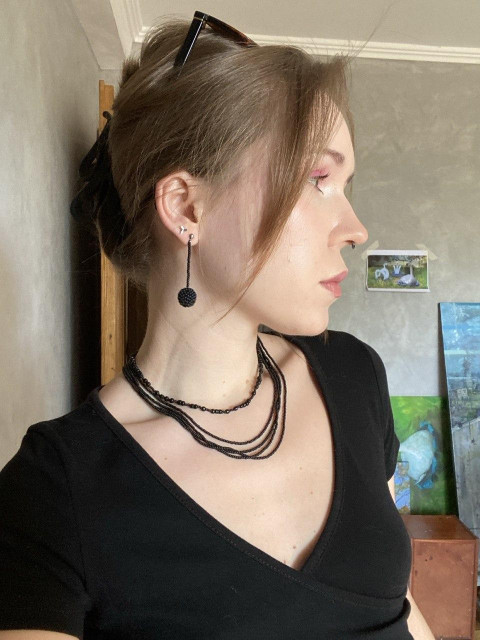 Side profile of a person wearing layered black beaded necklaces and long beaded earrings, dressed in a black top indoors.
