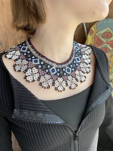 Close-up of a person wearing a detailed handmade beaded collar necklace with geometric floral patterns in black, white, blue, and soft pink tones.
