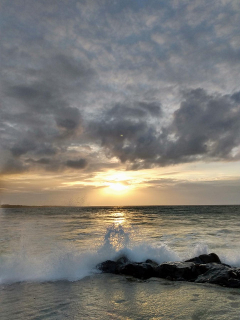 Under a lightly clouded evening sky, the sun sets over the sea on the horizon, its rays glowing golden-yellow. In the foreground, a wave with white foam crashes against a row of low stones jutting out into the picture from the right, along the flat, smooth sandy beach. 

Unter einem locker bewölkten Abendhimmel geht die Sonne am Horizont goldgelb über dem Meer unter. Im Vordergrund kracht eine Welle mit weißem Schaum an eine Reihe niedriger Steine, die von rechts am flachen glatten Sandstrand ins Bild ragen.