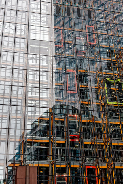 A vertical shot of a modern skyscraper’s glass facade. The windows create a dense grid that reflects an intricate network of internal or neighboring structures. Vibrant orange and red structural beams stand out sharply against the glass, mixed with subtle green accents and reflections of the sky, creating a complex, geometric pattern.