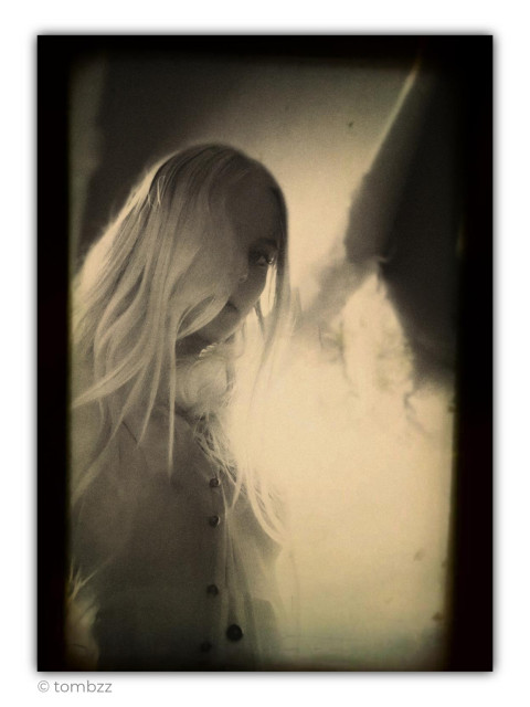 An artistic, monochromatic portrait in sepia tones, captured using analog technique with a characteristic vignette effect creating a natural dark frame around the edges. The model with long, light blonde hair stands with her back to a soft light source (softbox), creating a luminous aura around her silhouette. The exposure was consciously pushed to the edge of overexposure while maintaining subtle detail structure. Only half of the model's face is visible – one side emerges from the shadows, while the rest remains hidden behind strands of hair, giving the photograph a mysterious, ethereal atmosphere. She is wearing a light blouse with clearly defined dark buttons. The overall image is characterized by delicate grain and soft tonal transitions typical of classic fine art photography, building a mood of nostalgia and fleetingness.