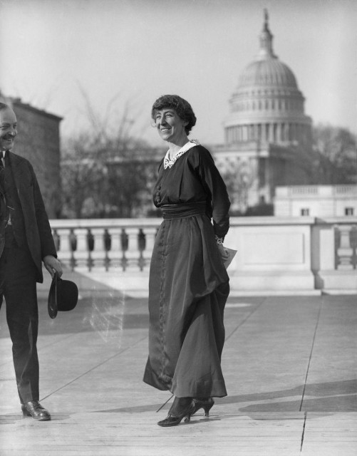 Jeanette Rankin standing in front of the dome of the US Capitol on a windy day. She is a white woman with dark hair.