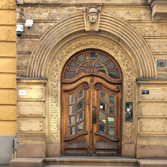 A majestic arched wooden entrance featuring highly decorative glass panes and scrollwork. The surrounding stone arch is intricately carved with repeating patterns and topped with a keystone depicting a man’s head wearing a winged helmet.