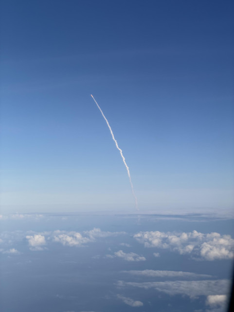 View out of the window of an aeroplane of the trail of smoke from the Artemis rocket against the blue sky, far above the clouds
