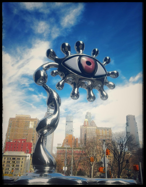 A color photo of a large sculpture made of a silver metal. On the left side are 3 large silver pieces in the shape of raindrops that balance on top of each other to make a pole. Coming out to the side of the pole is a giant human eye with a brown iris. Sprouting from the top and bottom of the eye are bulbus shaped silver pieces, like modern eyelashes. It is sitting on a silver base that looks like melted liquid silver. behind it are the buildings that line 25th St. and 26th St. to the north of Madison Square, Park. On the left side is a red and some light brick colored buildings and on the right side some older buildings sit behind several trees and stop lights. Further in the back is a more modern high-rise. The sky is very blue, streaked with white clouds.