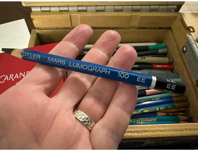 A hand holding up a Mars Lumograph 100EE pencil. It has a blue barrell and the lead is thick and dark.