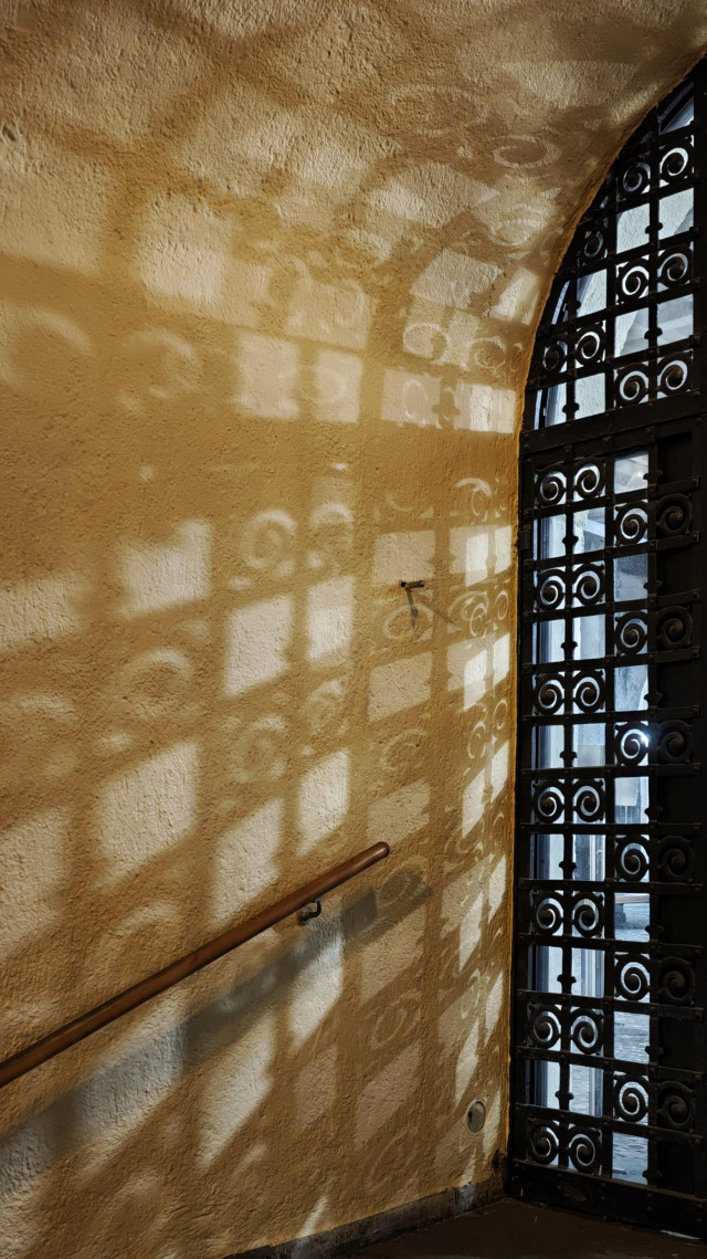 Das Foto zeigt eine große, mit kunstvollem, schwarzem, schmiedeeisernem Ziergitter versehene Festverglasung, die ein Seitenteil eines Eingangs mit einem halbkreisförmigen Oberlicht ist. Durch das Glas und das Gitter (schmale Streifen als quadratisches Gittermuster, bei dem immer zwei Quadrate nebeneinander einen Schnörkel haben und so eine Art 2x1-Schachbrettmuster bilden) fällt Licht, das ein komplexes Muster aus Schatten und Licht auf die angrenzende Wand wirft. Die Wand ist gelblich-beige und hat eine raue Struktur. An der Wand ist ein Handlauf aus Holz angebracht.