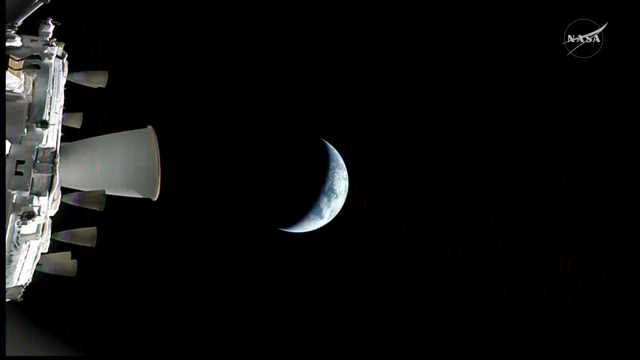 Still from webcast of the view from a camera on the Orion spacecraft, with a side view of the bottom of the spacecraft with thrusters on the left and a blue crescent earth in the center.