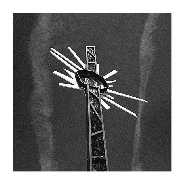 A stylized cross-like sculpture with radiating beams rises against a sky streaked with thin clouds.
