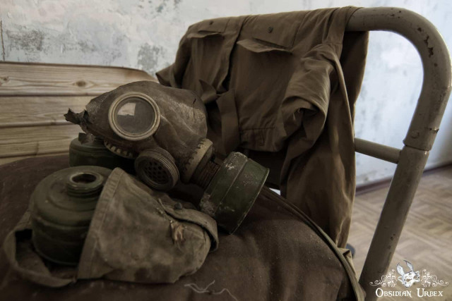 An old, dirty Soviet-era gas mask with a filter and a military jacket rest on a metal frame in an abandoned building.