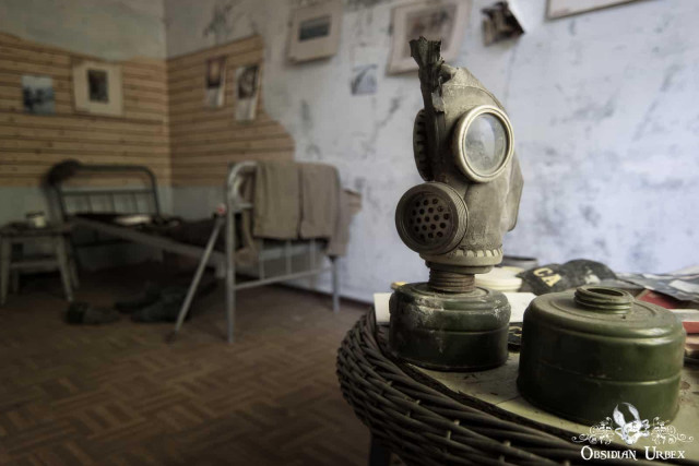 A weathered gas mask and its filters rest on a wicker surface, with a blurry, abandoned room