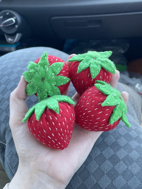 A photograph of a hand holding 4 hand-sewn felt strawberries. They’re red with green leaves and white rows of stitches running up and down the strawberries like seeds.