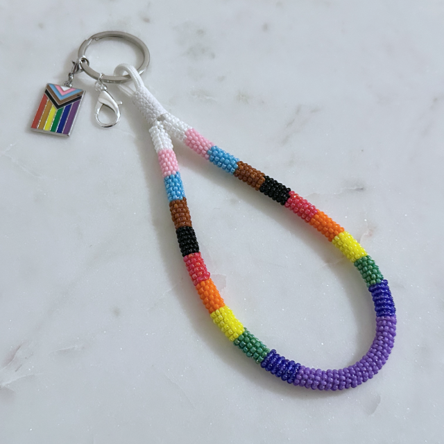 A bead wrapped wristlet keychain in the progress pride flag colors, with a silver ring, a lobster clasp clip, and a progress pride flag charm
