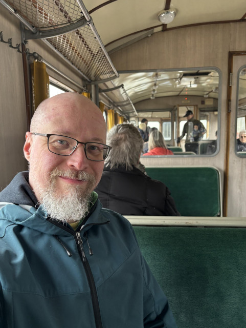 Selfie, jossa istun lättähatun vihreällä plyysipenkillä.