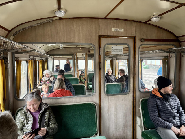 Lättähattujunan matkustamoa. Ihmisiä istuu vihreillä plyysipenkeillä. Kaarevakattoisen vaunun väliseinässä vaaleanruskea puupanelointi.