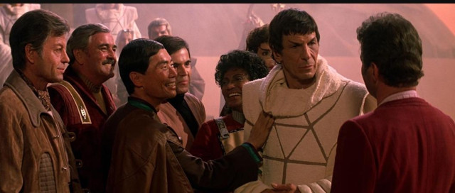 Star Trek The Search for Spock scene. Our Lord Spock is pictured in a white robe. He stands surrounded by his shipmates who are all older here than they were in the TV show. Sulu is smiling and touching Spock's chest for some reason. Uhura, Chekov, Scotty, and Bones stand close and all seem thrilled to see him, but are otherwise practicing self control and not feeling his chest. 

Despite being surrounded by friends, Spock is staring only at Kirk, for obvious reasons. 