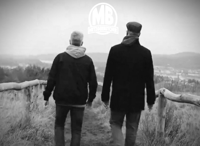 Zwei Männer gehen einen Weg von einem Hügel hinab. Sie sind von hinten zu sehen, im Hintergrund sieht man einen Fluss, einen Teil des Siebengebirges und die Stadt Bonn.