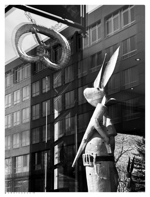 Aufnahme in schwarzweiß, im Schaufenster einer Bäckerei steht ein hoher handgefertigter Osterhase aus Holz, dieser hat eine Mohrrübe in der Hand und hat schöne lange gerade Ohren, in der Scheibe spiegelt sich ein großes Brezel und ein eckiges großes Gebäude mit vielen Fenstern  