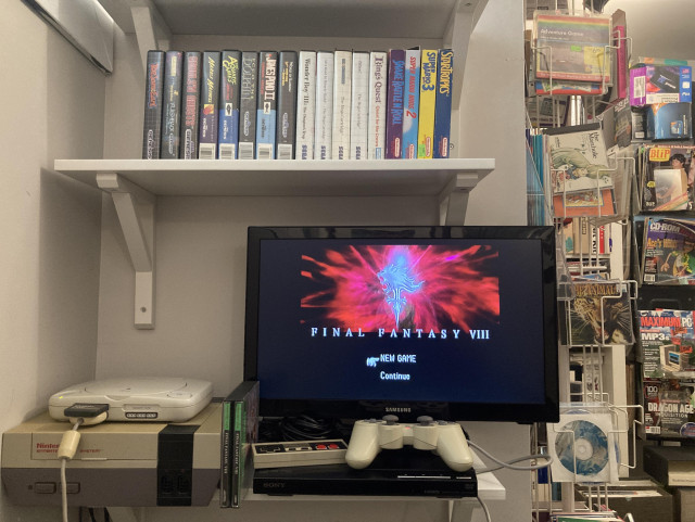 Two wall shelves hold a PSone console, Nintendo Entertainment System, and a small LCD tv. Final Fantasy VIII's splash screen menu is showing on the TV, with NEW GAME selected.