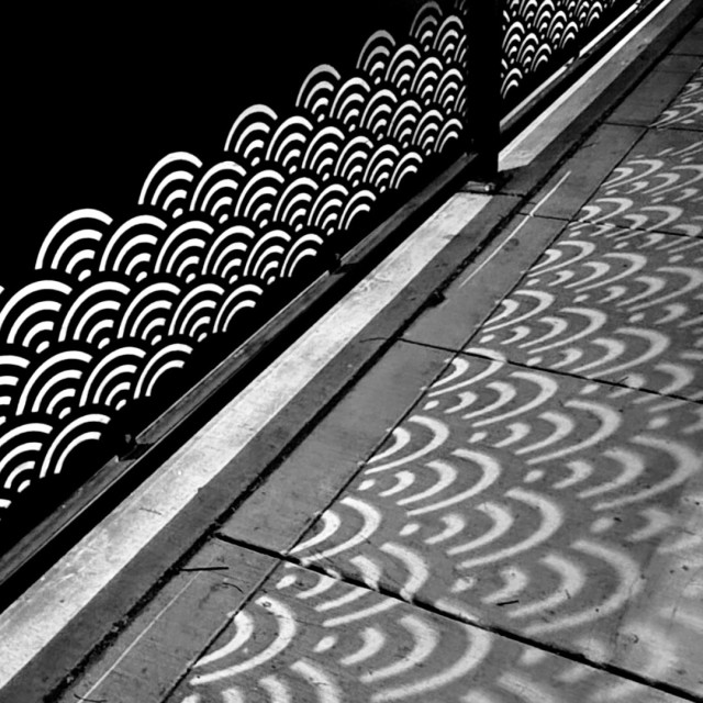 Black and white photo of a metal railing with a scalloped design casting shadows on a nearby sidewalk