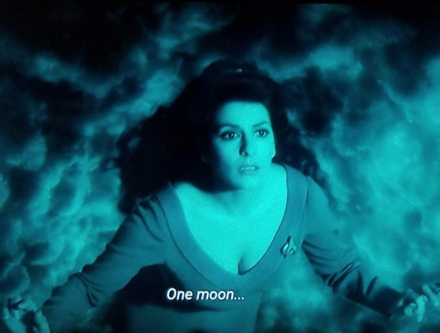 Star Trek The Next Generation scene. Deanna Troi is floating in space in a swirl of green dreamy clouds or maybe a nebular. She's looking up and we're looking down at her. Her arms are open and she looks shooketh or worried or entranced otherwise. Closed caption reads, "One moon..."