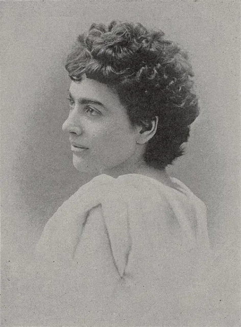 Image is of a black & white formal photograph portrait on a young Caucasian woman.  Her body is turned away from the camera, and her face is turned back forty-five degrees (so she is still forty-five degree from facing front).

She has thick, curly dark hair, dark eyebrows, and apparently dark eyes.  A strong-looking woman, but with soft, not angular, features.  She is wearing a white dress or blouse of some kind, but all the focus is on her head & shoulders and we cannot see the detail of of anything else.