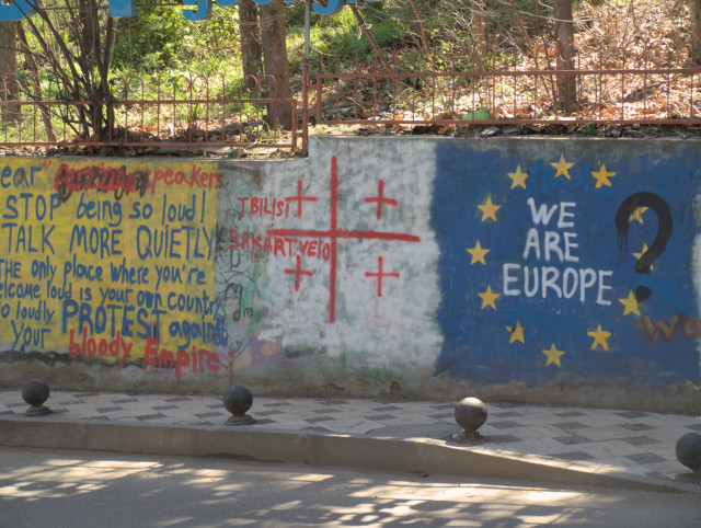 Political painting on a wall with message:
Dear Russian speaker Stop being so loud!
Talk more quietly.
The only place where you're 
welcome loud is your own country
to loudly protest against 
your bloody Empire!
Georgian flag.
EU flag with the words:
We are europe
Someone has put a question mark there.