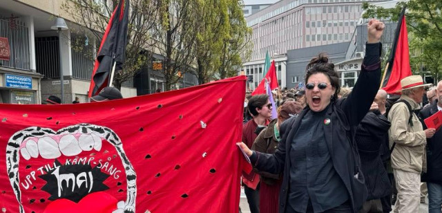 Foto från 1 maj 2025, Göteborg : Svartklädd demonstrant med höjd knuten näve står invid röd banderoll föreställande ett sjungande gap, och med texten "Upp till kamp-sång".