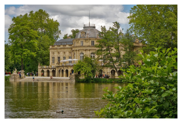 The photo shows Lakeside Palace Monrepos in Ludwigsburg, Germany. An elegant Rococo-style palace set beside a lake. The building has a light yellow, classical façade with many large windows, arched openings at ground level, and a prominent central dome. The palace is surrounded by a lush green park. Tall trees frame the scene on both sides, and branches with dense leaves partly cover the right side of the image. In the foreground, the calm lake stretches across the lower half of the picture, reflecting parts of the palace and the surrounding greenery. A few ducks are swimming on the water, and several people can be seen near the shoreline and terrace area. The sky is mostly cloudy, which gives the scene soft, even lighting. Overall, the image conveys a peaceful, scenic atmosphere and presents the palace as a refined historic building in a natural park setting.