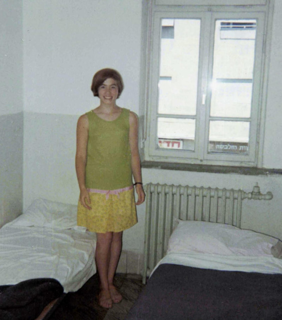 A teenage girl in a sleeveless dress, standing next to two basic beds in a cheap hotel room. She has a big smile. 