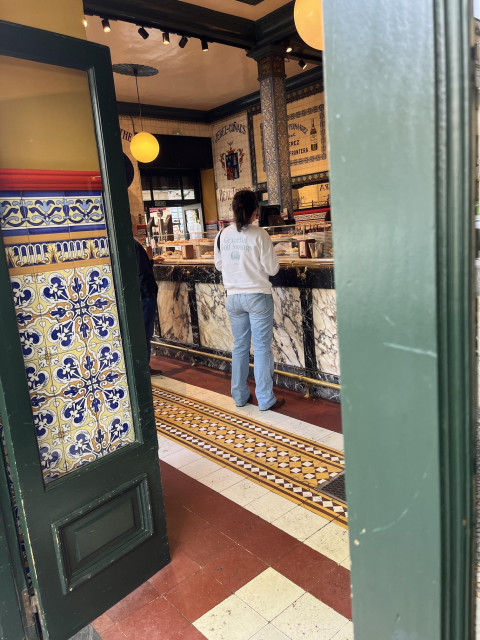 View through door of bar/café dating back at least to the early 1900s. The doors are painted green and have large windows in them. But they're fully open. The walls are mostly covered by decorative tiles in various Victorian-looking patterns (floral, geometric). The floor tiles are matte as opposed to the glossy look of the wall tiles. They're solid colors instead of printed or painted like the wall tiles. However, the solid color pieces are laid out in geometric patterns. The bar is black marble with white veins around the edges and white marble with black veins in rectangular panels. The columns up to the high ceilings are also covered with tile and the walls opposite have some tiles with words baked into the tiles. A woman wearing pants and a sweatshirt that says "Graceful Golf Swings" stands at the bar. Another customer stands at the bar almost out of the door's frame.