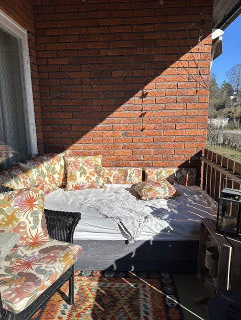 Ett soligt hörn på en indragen balkong på en röd tegelvilla. I bortre hörnet en säng med kuddar runt omkring.
