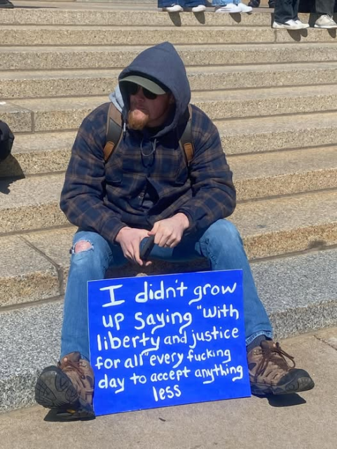 Photo of a man sitting on some large steps (as if in front of a government building) with a sign that reads, "I didn't grow up saying 'With liberty and justice for all' every fucking day to accept anything less."