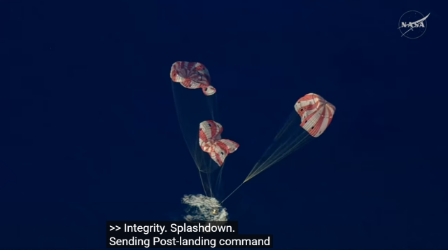 A captured still from the linked live NASA broadcast showing the Orion spacecraft capsule, seen from above, landing in ocean water with three red-and-white parachutes still mid-air, descending down.

On-screen caption reads: "Integrity. Splashdown. Sending Post-landing command."