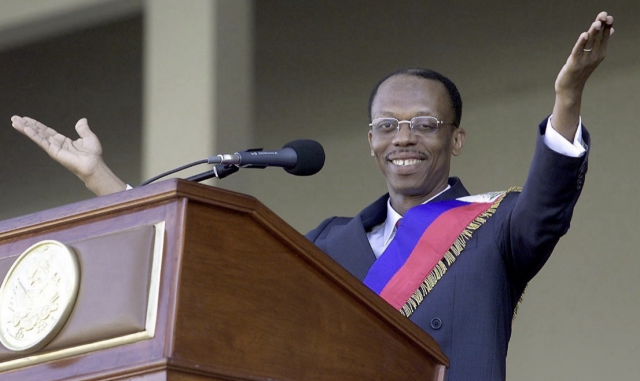 Jean-Bertrand Aristide, wearing a presidential sash, raises both arms while speaking at a podium during his inauguration ceremony in Port-au-Prince in 1991.