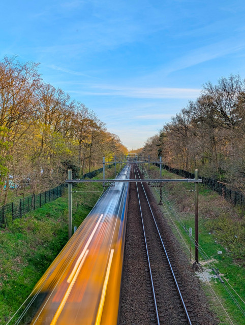 Two parallel sets of tracks run vertically through the frame. The right track is empty and in sharp focus, showing the texture of the dark gravel (ballast) and the glint of the metal rails. On the left track, a train is blurred into long, glowing streaks of yellow, orange, and white light.